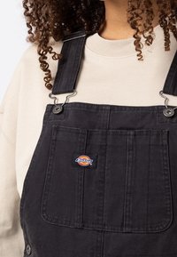 Zwarte tuinbroek van duurzaam stof met metalen clips en een voorzak. Heeft een Dickies-logo op de voorkant. Lichtbeige shirt eronder.