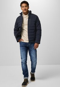 Mörkblå puffjacka med dragkedja och fickor över en beige hoodie, kombinerad med blå jeans och svarta snörstövlar med strukturerad sula.