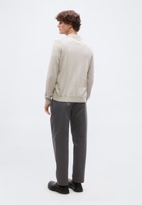 Νέος άνδρας με σγουρά μαλλιά, που στέκεται και κοιτάει προς τα αριστερά, φορά ανοιχτό μπεζ μακρυμάνικο πόλο, σκούρα γκρι παντελόνια και μαύρα παπούτσια σε λευκό φόντο.