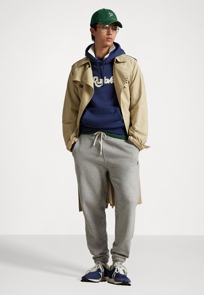 Palton bej de tip trench, hanorac albastru cu logo, pantaloni de trening gri și adidași albaștri. O șapcă verde și ochelarii completează ținuta casual.
