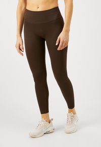 Barna bordás leggings magas derékkal, testhezálló kialakítással és varrás nélküli szerkezettel, világos színű sportcipővel párosítva.