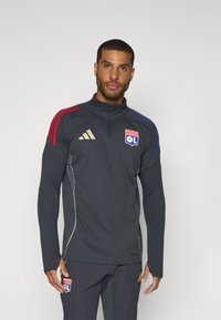 Mørkegrå atletisk jakke med kvarte zip, røde og blå skulderstriber, gyldne detaljer og logoen fra Olympique Lyonnais på brystet.
