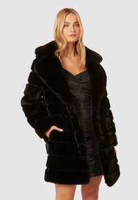 Cappotto in pelliccia sintetica nera con ampio colletto e design trapuntato, dotato di due tasche frontali e una texture morbida, indossato sopra un vestito scuro e luccicante.