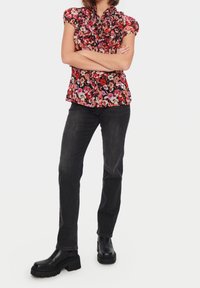 Blouse fleurie à col à volants, manches courtes bouffantes en rouge, rose et noir. Associée à un jean denim foncé et des bottes noires épaisses.