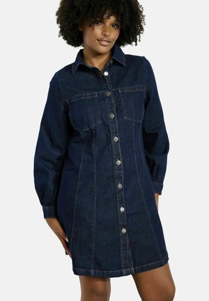 Woman with curly hair wearing a dark blue denim button-up dress with long sleeves, front pockets, and a collar, standing against a white background.