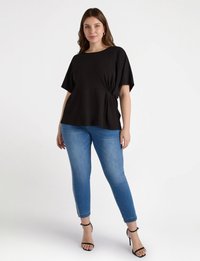 Schwarzes kurzärmliges Oberteil mit gerafftem Bund, kombiniert mit schmal geschnittenen blauen Jeans, die knöchellang sind und silberne Details an den Seiten haben.