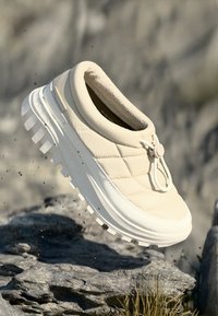 Beige tøfler med quiltet syntetisk overdel, elastisk snor med lås, og hvit gummisåle med tykk, teksturert såle.