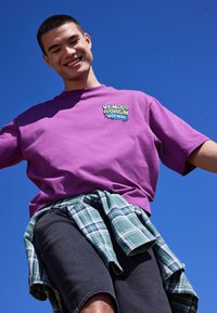 Jeune homme souriant, portant un t-shirt violet "Venice Beach Hip Hop" et une chemise à carreaux nouée autour de la taille, devant un ciel bleu clair.