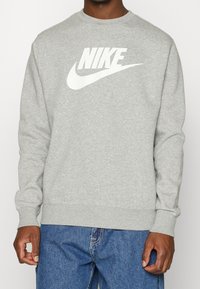 Grå sweatshirt med rund halsringning, ribbade ärmslut och nederkant. Har en stor vit Nike-logotyp på framsidan. Tyget verkar mjukt med en slät textur.