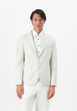 Man wearing a cream pinstripe blazer over a white shirt with black buttons and white trousers, standing against a plain white background.