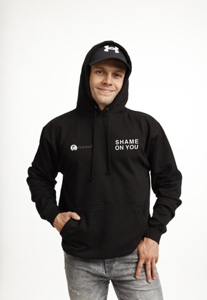 Ung mand iført en sort hættetrøje med teksten "SHAME ON YOU" og en sort Under Armour-kasket, smilende med den ene hånd i lommen og den anden på låret.