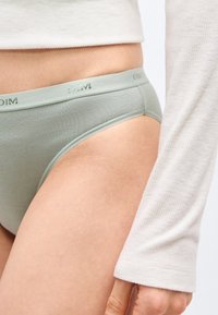 Κοντινό πλάνο ενός ατόμου που φοράει βαμβακερά εσώρουχα ανοιχτού πράσινου χρώματος με το λογότυπο "DIM" στη μέση και ένα λευκό μακρυμάνικο μπλουζάκι.