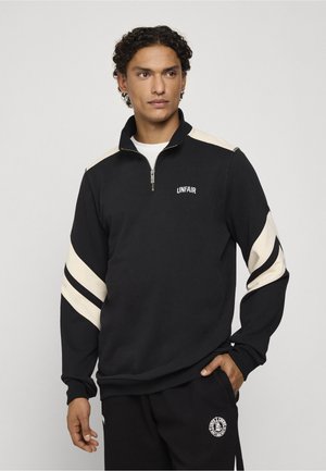 Homme portant un sweat à capuche noir zippé avec des bandes blanches sur les manches et le texte "UNFAIR" sur la poitrine, debout avec une main dans la poche.