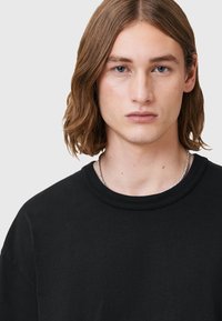 T-shirt nera in cotone con scollo rotondo e maniche corte. Presenta una texture uniforme. Il modello indossa una collana in argento.