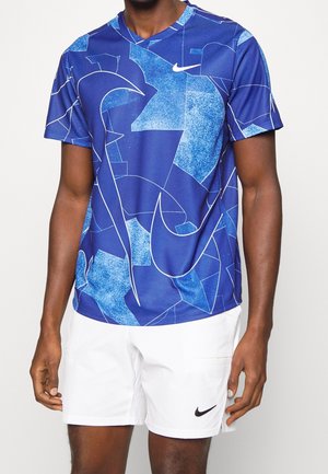Mann trägt ein blaues, abstrakt gemustertes kurzärmeliges Hemd und weiße Shorts, beide mit schwarz-weißen Nike-Logos.