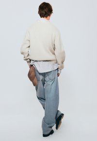 Suéter oversized color crema, superpuesto a una camisa a cuadros. Jeans baggy de color azul claro, zapatos negros y una gorra marrón metida en la cintura.