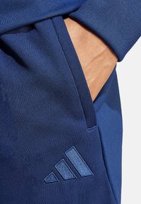 Marinefarbene Sporthosen mit glatter Textur, ausgestattet mit einer Seitentasche und einem bestickten Drei-Streifen-Logo in einem helleren Blauton.