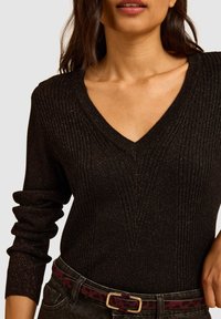 Zwarte gebreide trui met V-hals, voorzien van verticale ribbels en subtiele glitter. Draagt met hooggerokte jeans en een gemusterde riem.