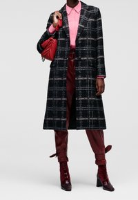 Soprabito a quadri in bianco e nero con silhouette lunga, indossato sopra una camicia rosa e pantaloni in ecopelle bordeaux. Borsa rossa e stivali coordinati.