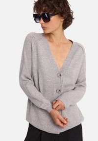 Jasnoszary, dziergany kardigan z głębokim dekoltem w kształcie litery V, dwoma guzikami oraz prążkowanymi mankietami i dołem. Gładka faktura, luźny krój, zestawiony z czarnymi spodniami.