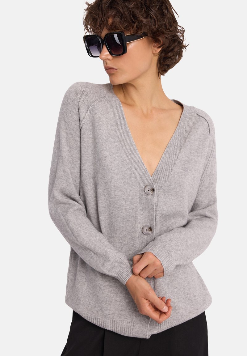 Jasnoszary, dziergany kardigan z głębokim dekoltem w kształcie litery V, dwoma guzikami oraz prążkowanymi mankietami i dołem. Gładka faktura, luźny krój, zestawiony z czarnymi spodniami.