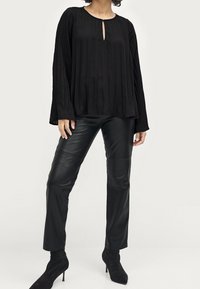 Femme portant un blouse plissée noire à manches longues avec encolure en trou de serrure, un pantalon en cuir noir et des bottines à talons noires, se tenant contre un fond blanc.