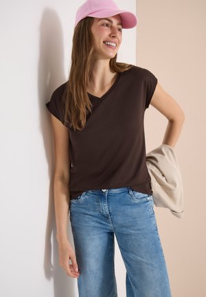 Ung kvinde smilende, iført en pink kasket, sort ærmeløs top og blå jeans, holder en beige jakke mod en tofarvet væg.