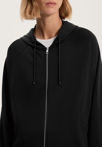 Črna zip-up kapuca iz gladkega materiala, ki ima kapuco, vrvico in celotno zadrgo z minimalističnim dizajnom.