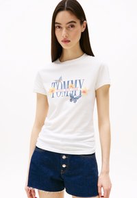 Witte katoenen t-shirt met grafische print met "TOMMY JEANS NEW YORK," bloem- en vlinderaccenten. In combinatie met donkere denim shorts.