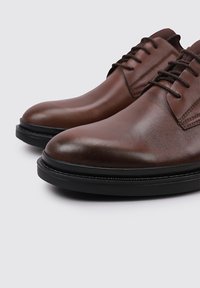 Zapatos de vestir de cuero marrón con textura suave, puntera redonda y suela de goma negra. Presentan cordones tradicionales y detalles de costura sutiles.