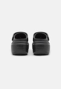 Des Crocs noirs vus de l'arrière, avec une texture à pois sur la semelle, une sangle arrière et un design ouvert. Le nom de la marque est visible sur le talon.