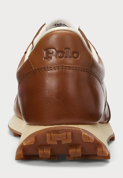Sapatilha de couro castanho com detalhes brancos, apresentando uma superfície texturizada, logo "Polo" em relevo e sola de borracha com padrões de aderência.