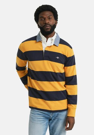 Man in gele en marineblauwe gestreepte rugbyshirt met een lichtblauwe kraag en lichtblauwe jeans, staand met één hand achter zijn rug.