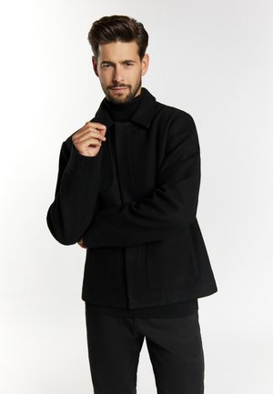 Schwarze Wollmischjacke mit hohem Kragen, Fronttaschen und strukturierter Silhouette. Das Model posiert mit neutralem Ausdruck.