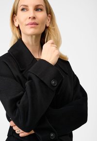 Sort uldblandingsfrakke med store revers og knapmansjetter. Har en blød tekstur og et struktureret design, der understreger varme og dækning.