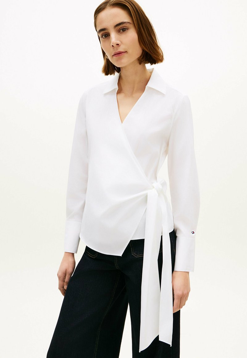Femme portant un chemisier blanc à nouer avec des manches longues à poignets larges et un nœud sur le côté, associé à un pantalon large taille haute sombre sur un fond uni.
