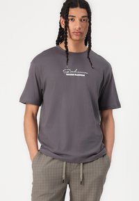 Junger Mann mit langen geflochtenen Haaren, der ein dunkelgraues T-Shirt mit dem Aufdruck "Sakura Blossom" und hellgraue karierte Hosen trägt, die Hände in den Taschen.