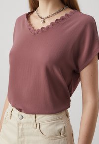 Kortärmad mauve-blus med en texturerad yta och en scalloped V-ringning. Tillsammans med ljusa högmidjade jeans.