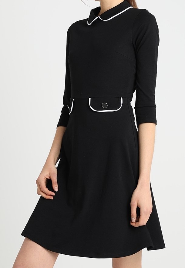 Femme portant une robe noire avec des manches trois-quarts, un liseré blanc sur le col et les poches, et un détail bouton sur la poche à la taille.
