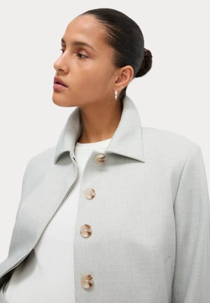 Mladá žena s uhlazenými vlasy, která má na sobě stříbrné kruhové náušnice a světle šedý kabát na knoflíky přes bílý top, dívá se doleva.