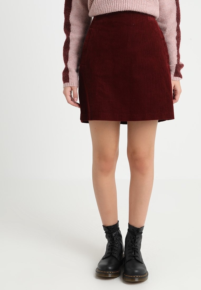 Personne portant une jupe en velours côtelé bordeaux, un pull rose et bordeaux, et des bottines noires à lacets, debout sur un fond blanc.