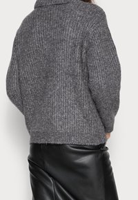 Person som bär en grå stickad tröja och en svart läderkjol, sedd bakifrån mot en enhetlig bakgrund.