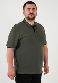 Tmavozelený polo tričko s čiernym golierom, krátkymi rukávmi a tromi gombíkmi. Mäkkučký materiál, rovný strih, s malým logom na hrudi.