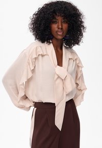 Jasnoróżowa bluzka z falbaniastymi rękawami i związanym detalem przy kołnierzyku, zestawiona z ciemnobrązowymi spodniami o wysokim stanie z kremowymi paskami po bokach.