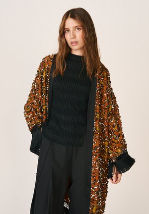 Femme aux longs cheveux ondulés portant un haut côtelé noir, un pantalon noir à jambes larges et un manteau ouvert orange orné avec des poignets à franges noires.