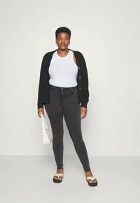 Joven mujer vestida con una camiseta de tirantes blanca, cárdigan negro, jeans ajustados oscuros y sandalias blancas, sosteniendo una bolsa de tela blanca, de pie contra una pared lisa.