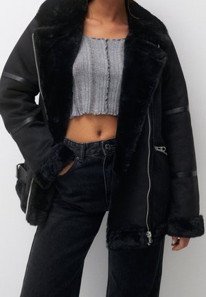 Femme portant un haut court gris à côtes, un jean noir taille haute, et une veste en peau lainée noire ouverte avec des fermetures éclair et une bordure en fourrure.