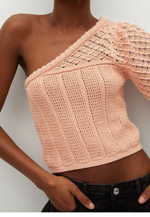 Brzoskwiniowy dzianinowy top na jedno ramię z fakturowanymi wzorami, noszony z czarnymi spodniami, ręka spoczywająca w kieszeni, na jednolitym tle.