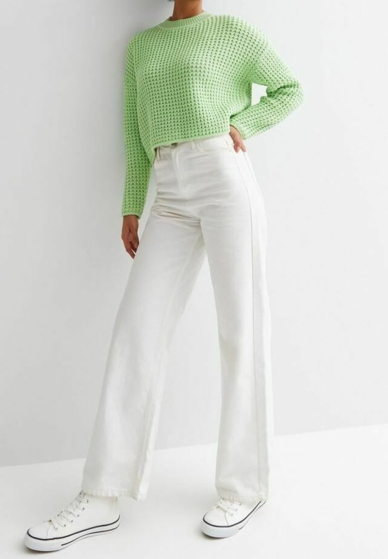 Personne portant un pull texturé vert clair, un pantalon large blanc taille haute et des baskets blanches avec des bordures noires, debout devant un fond blanc.