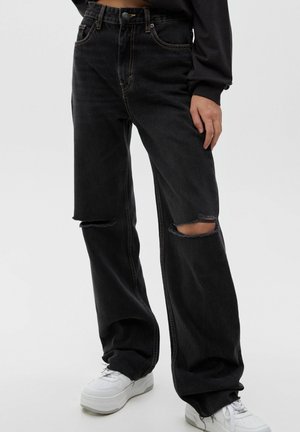 Jeans de mezclilla negra con un corte holgado, que presentan grandes desgarros en las rodillas y costuras naranjas en contraste. Combinados con zapatillas blancas.